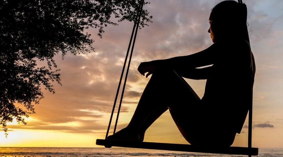 A serene sunset silhouette of a woman sitting on a swing by the seashore, evoking tranquility and reflection.